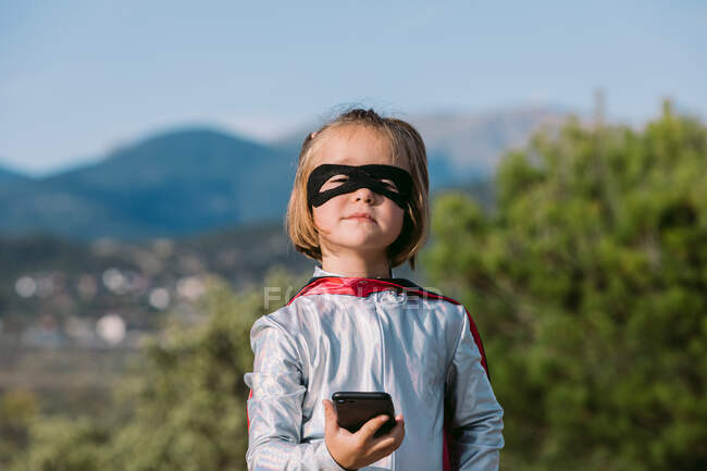 Auto assuré fille en masque oculaire costume de super-héros avec cape navigation sur téléphone portable — Photo de stock