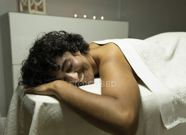 Jovem senhora étnica positiva sob toalha deitada no sofá com olhos fechados no salão de spa luz — Fotografia de Stock