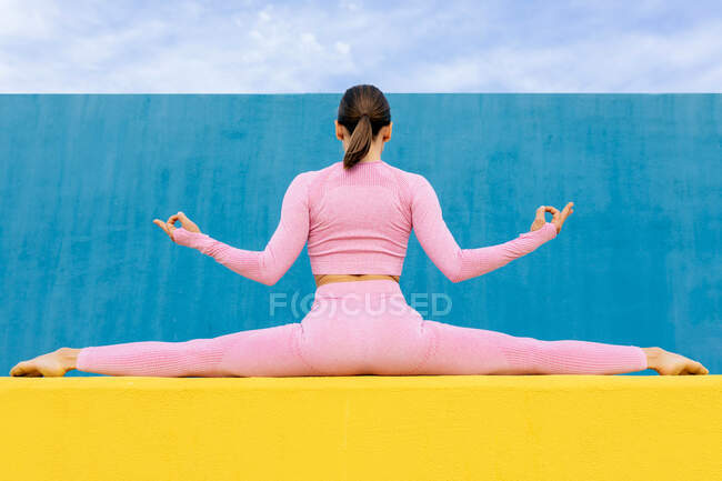 Back view of slim female sitting in split doing gyan mudra while sitting on high yellow wall on blue background — Stock Photo