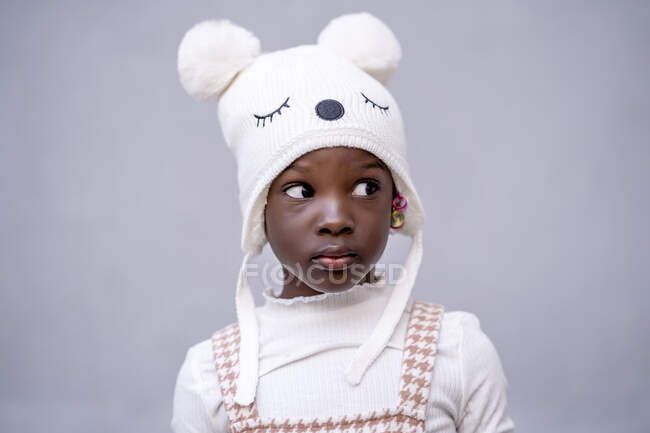 Petite fille afro-américaine sérieuse en vêtements élégants et chapeau drôle debout regardant loin sur fond gris — Photo de stock