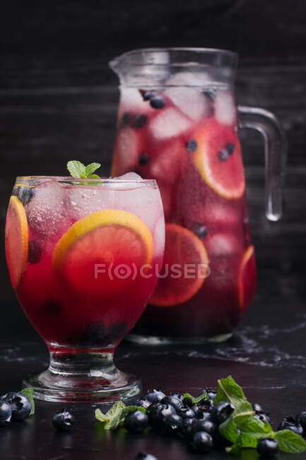 Glass and jug with refreshing cold lemonade with fresh blueberries and lemon slices placed on dark table — Stock Photo