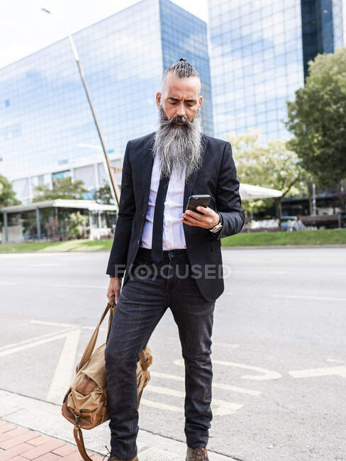 Selbstbewusster bärtiger Mann im edlen Anzug surft auf dem Smartphone, während er am Straßenrand auf einer Straße mit modernen Gebäuden in der Stadt steht — Stockfoto