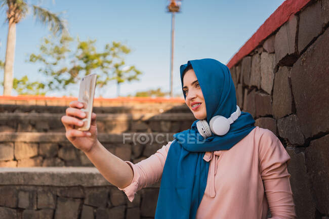 Mulher muçulmana encantadora em lenço de cabeça tradicional em pé perto da parede de pedra e tomando auto-tiro no smartphone no dia ensolarado — Fotografia de Stock