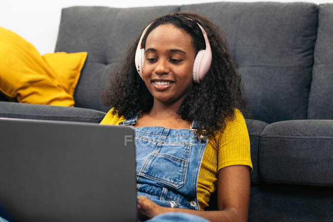 Mujer negra sonriente en overoles de mezclilla con auriculares sentados en el suelo cerca del sofá y utilizando el ordenador portátil en casa - foto de stock