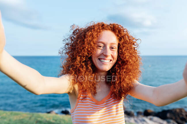 Glücklich gelockte Frau, die die Arme ausstreckt und in die Kamera schaut, während sie auf einem Smartphone Selbstporträts an der Küste des Meeres macht — Stockfoto