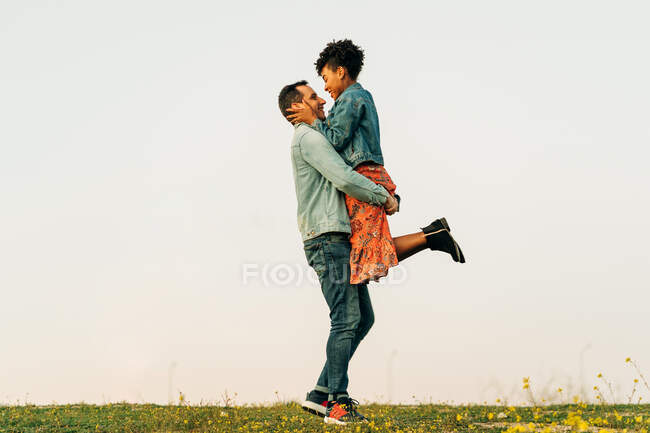 Vue latérale d'un jeune homme romantique en vêtements décontractés embrassant et portant une petite amie ethnique souriante sur une prairie herbeuse verte sous le coucher du soleil — Photo de stock