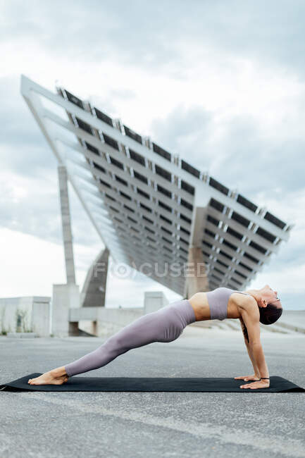 Full body side view of sportive female in activewear practicing Purvottanasana posture on mat in city near modern solar panel — Stock Photo