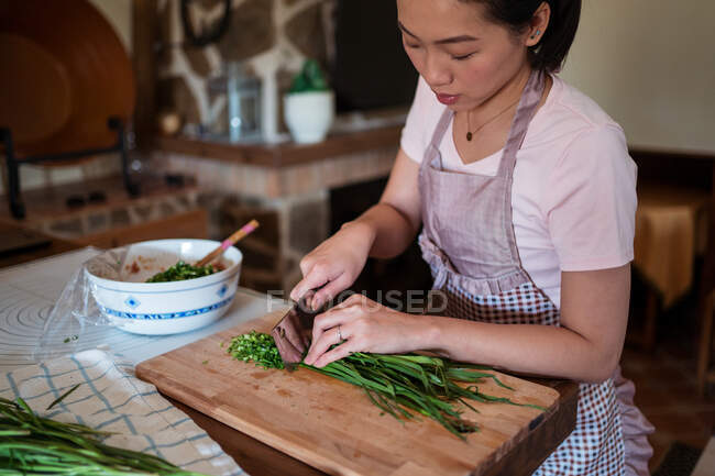 De cima de mulher cortando ervas verdes frescas na tábua de corte de madeira enquanto prepara o jantar na cozinha — Fotografia de Stock