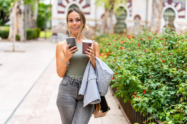 Fröhliche Frau mit Kaffee zum Mitnehmen SMS auf dem Handy und Blick in die Kamera, während sie auf der Straße in der Nähe von grünen Büschen auf verschwommenem Hintergrund steht — Stockfoto