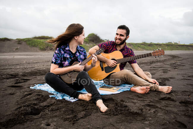 Voller Länge lächelnder Mann, der Akustikgitarre spielt, mit positiver Freundin, die Ukulele spielt, während sie an der Sandküste in der Natur bei bewölktem Tag sitzt — Stockfoto