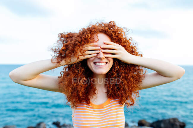 Unrecognizable female with long ginger curls covering face with hands while standing on coast of blue sea — Stock Photo