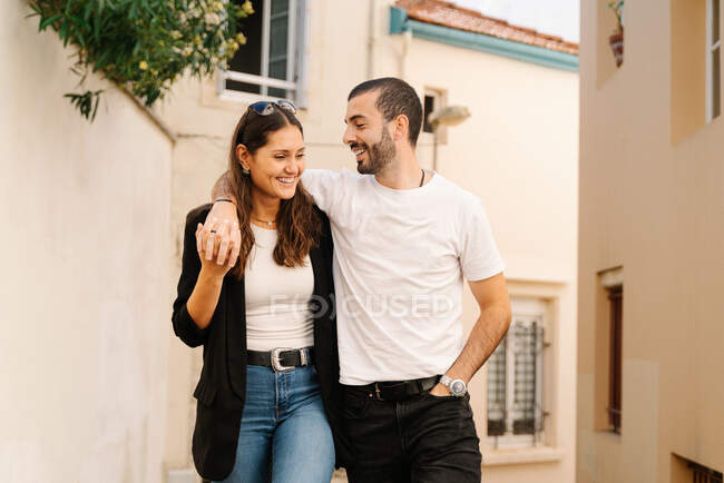 Eccitato giovane elegante coppia ispanica coccole e sorridente mentre si cammina sulla stretta strada tra l'edificio residenziale invecchiato nella giornata di sole — Foto stock