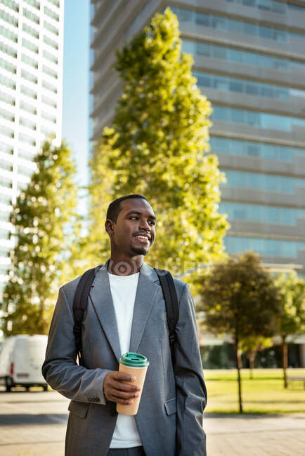Позитивный афроамериканец с рюкзаком, стоящим с одноразовой чашкой кофе и смотрящим в сторону с зубастой улыбкой — стоковое фото