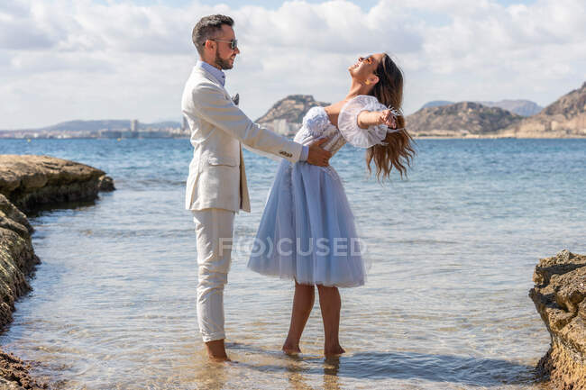 Side view of groom hugging bride with spread arms while standing on shore near rippling sea during wedding celebration in nature on summer day — Stock Photo