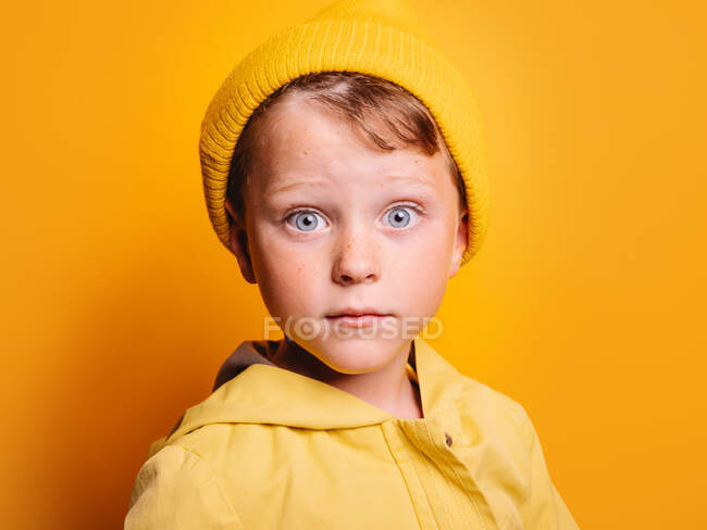 Amazed little boy with blue eyes in bright yellow autumn jacket and beanie hat looking at camera against yellow background in studio — Stock Photo
