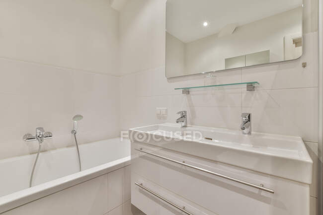 Armario con lavabo y espejo situado cerca de la bañera en el moderno baño de luz - foto de stock