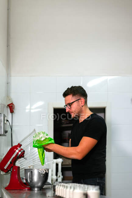Vista lateral de un joven panadero masculino tatuado concentrado en ropa casual y anteojos llenando la bolsa de pastelería con crema mientras prepara deliciosa torta en la cocina. - foto de stock