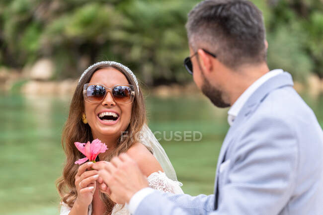 Positive married couple in wedding outfit and sunglasses sitting on stone stair near lake and green palms and plants while looking at each other and giving flower — Stock Photo