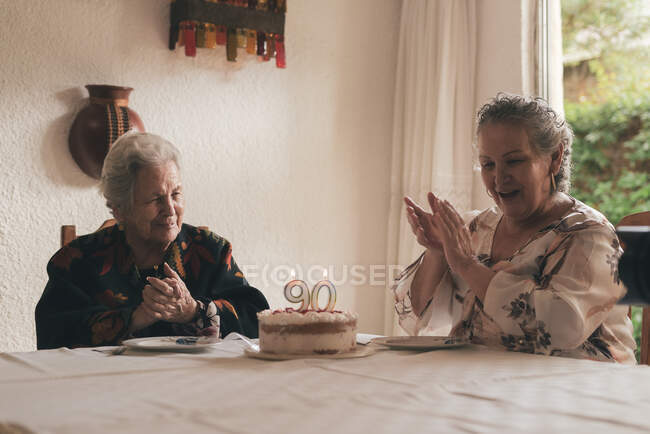 Ältere Frau mit grauen Haaren und ältere Dame sitzen am Esstisch und feiern 90. Geburtstag mit leckerem Kuchen mit Kerzen — Stockfoto
