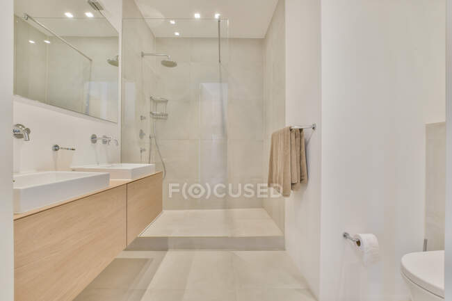 Lavabos blancos en la pared con espejo situado cerca de cabina de ducha de vidrio en baño amplio y luminoso con toalla e iluminación brillante - foto de stock
