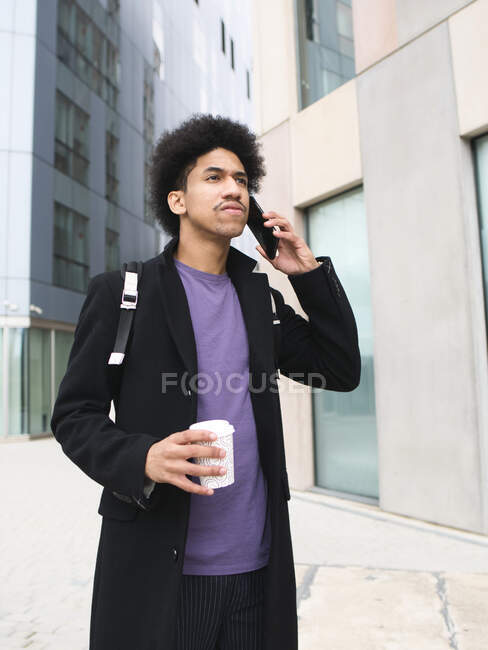 Serious young black male millennial with Afro hair in trendy outfit standing on city street with takeaway coffee and talking smartphone — Stock Photo
