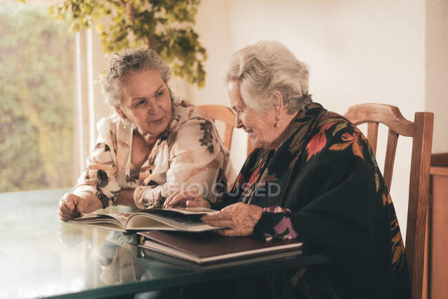 Elderly sisters inspecting pictures in photo album and discussing memories while sitting at table at home together — Stock Photo