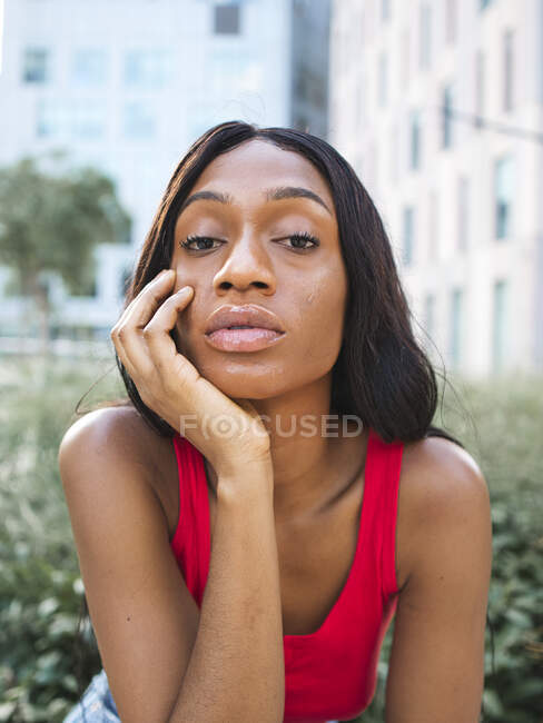 Mulher afro-americana confiante em cima e jeans olhando para a câmera enquanto sentado na cova de flores com plantas verdes na rua com edifícios modernos de vários andares — Fotografia de Stock