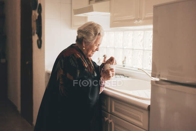 Vista lateral da fêmea idosa em xale quente com caneca em pé perto da pia branca na cozinha branca clara — Fotografia de Stock