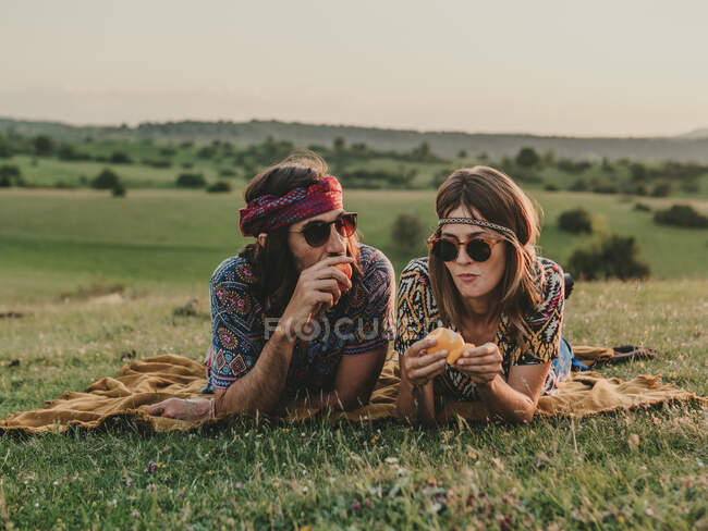 Пара хиппи в солнечных очках чистит и ест апельсин, пока лежит на травянистом лугу на природе на закате — стоковое фото