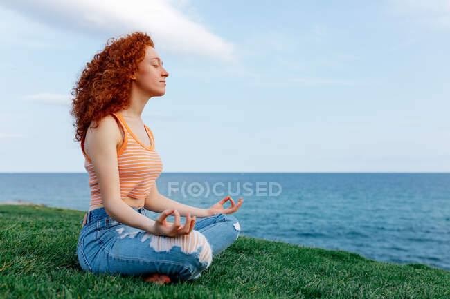 Side view of carefree female with long ginger hair in Padmasana pose with zen gesture on grassy hill on seashore — Stock Photo