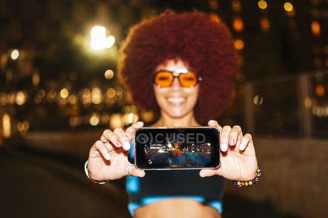 Mujer feliz con peinado afro tomando autorretrato en el teléfono inteligente mientras está de pie en la calle oscura con farolas sobre fondo borroso - foto de stock