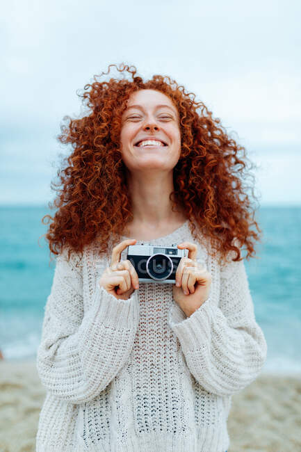 Mujer pelirroja positiva en suéter de punto de pie con los ojos cerrados mientras sostiene la cámara fotográfica retro en la costa del mar - foto de stock