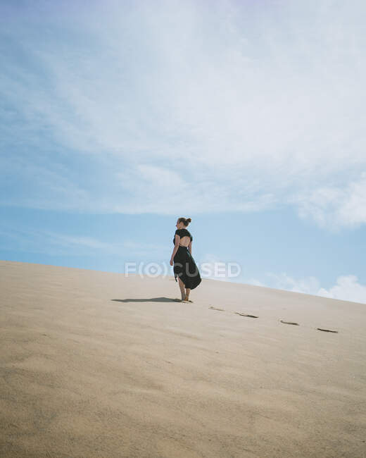 Vista trasera de cuerpo completo de una hembra descalza irreconocible en vestido paseando sobre una duna arenosa en el desierto - foto de stock