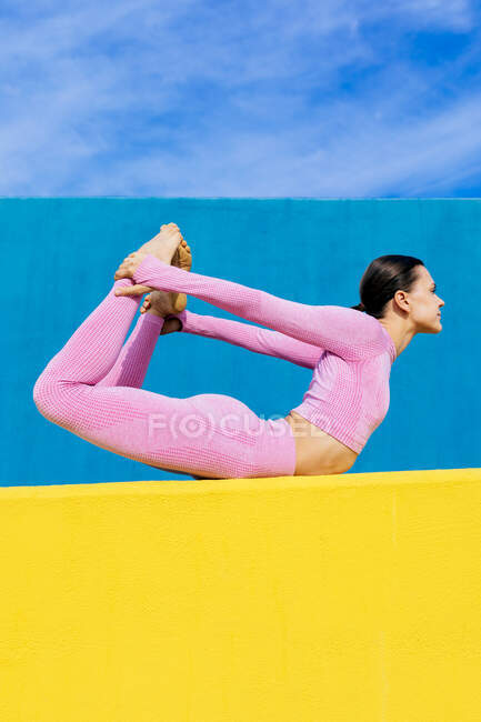 Vista lateral de cuerpo completo descalzo hembra haciendo pose de arco mientras entrena pose Dhanurasana en la pared amarilla sobre fondo azul - foto de stock