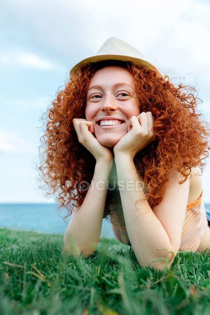 Happy curly redhead haired female with freckles lying on lawn looking away on coast of sea — Stock Photo