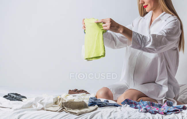 Беременная женщина с красными губами, ожидая ребенка складывая одежду детей, сидя на кровати в спальне — стоковое фото