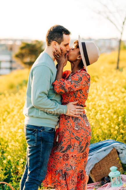 Vista lateral de la joven mujer afroamericana feliz con sombrero sonriendo mientras abraza y besa novio con los ojos cerrados de pie en el prado floreciente en el día soleado - foto de stock