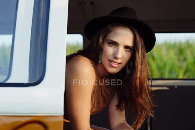 Симпатичная брюнетка в шляпе в винтажном фургоне и сидит на сиденье и смотрит в камеру в солнечный день — стоковое фото
