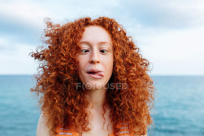 Juguetona hembra de pelo rizado mostrando la lengua y cruzando los ojos mientras hace mueca divertida en la costa de mar ondulante azul - foto de stock