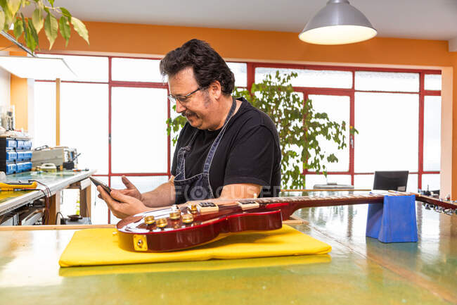 Positive adult male master in apron and eyeglasses while using mobile phone near table with various instruments and electric guitar near windows and plants in light workshop — Stock Photo