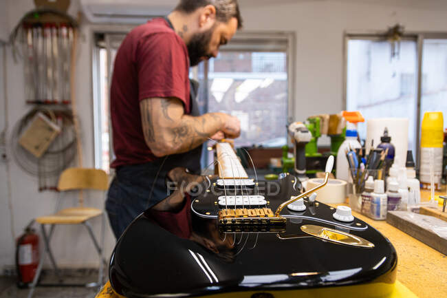 Side view of crop concentrated craftsman with tattoos standing and repairing electric guitar in workshop in daytime — Stock Photo
