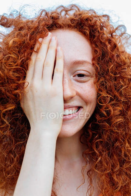 Jovem feliz encaracolado ruiva fêmea com olhos castanhos e sardas cobrindo o olho com a mão e olhando para a câmera — Fotografia de Stock