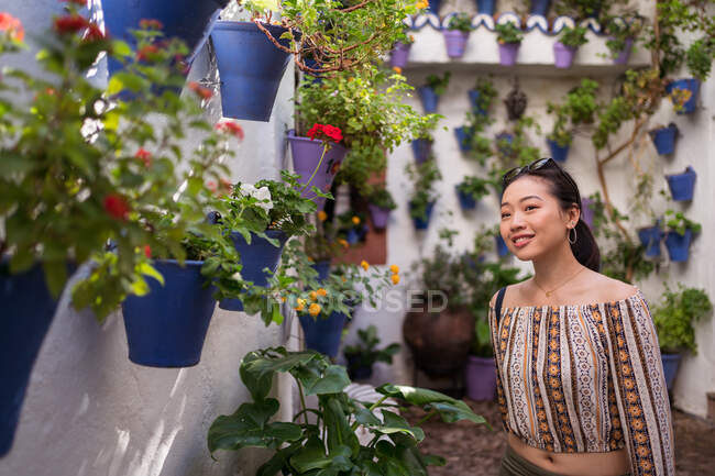 Positivo Asiático turista feminino em óculos de sol olhando para longe, enquanto de pé perto do edifício com flores coloridas florescendo em vasos de flores na cidade — Fotografia de Stock