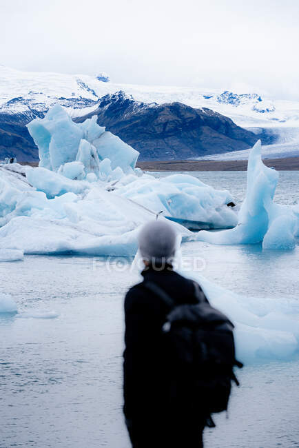 Rückenansicht eines anonymen Wanderers in warmer Oberbekleidung und Hut, der auf einem felsigen Hügel steht und die atemberaubende Landschaft des Jokulsarlon großen Gletschersees während einer Reise in Island bewundert — Stockfoto