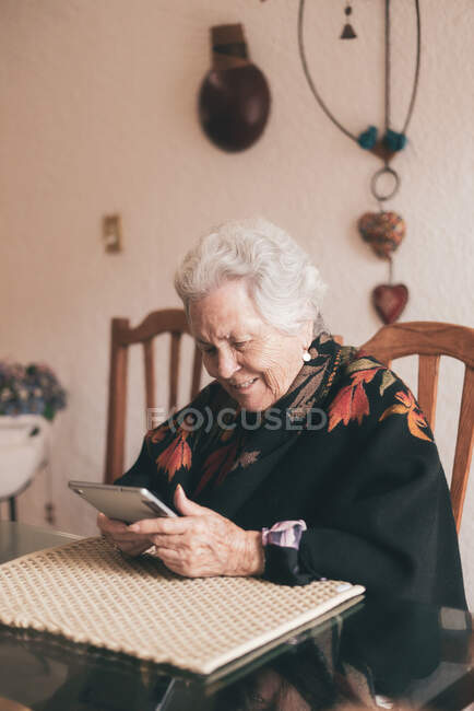 Улыбающаяся пожилая женщина в теплой одежде сидит за столом с табличкой и чашкой чая, глядя на экран — стоковое фото