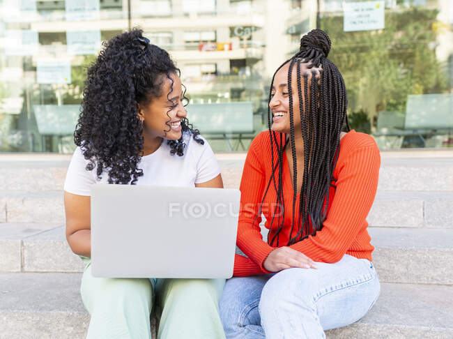 Allegro diverse amiche donne in abiti alla moda seduti sulla scala e guardando l'un l'altro durante la navigazione laptop sulla strada della città in giorno d'estate — Foto stock