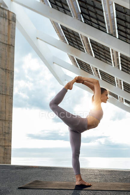 Ganzkörper aktive Frau in Sportbekleidung, die mit gebeugten Knien stehend auf einem Bein in der Nähe von Photovoltaikanlagen auf der Straße steht — Stockfoto