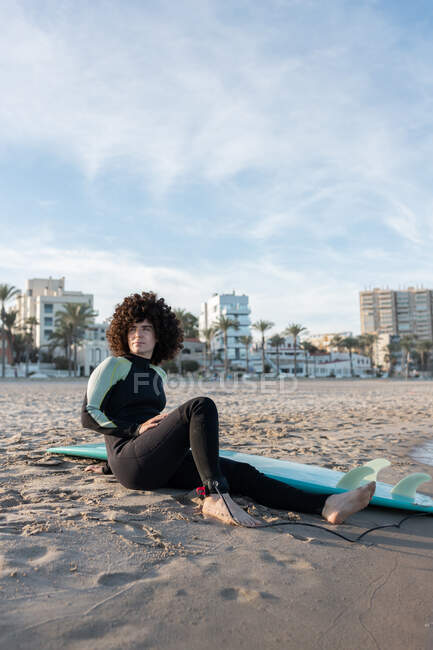 Вид сбоку: полное тело женщины-серфингистки в гидрокостюме, сидящей на песчаном пляже рядом с морем — стоковое фото