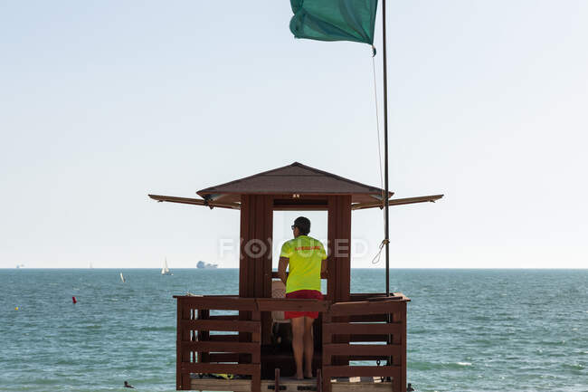 Vue arrière complète du corps d'un sauveteur masculin méconnaissable contrôlant la sécurité en mer depuis une tour de sauvetage en bois — Photo de stock