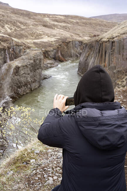 Visão traseira do viajante irreconhecível em roupas casuais em pé no topo do penhasco e tirar foto do rio no desfiladeiro Studlagil na Islândia à luz do dia — Fotografia de Stock